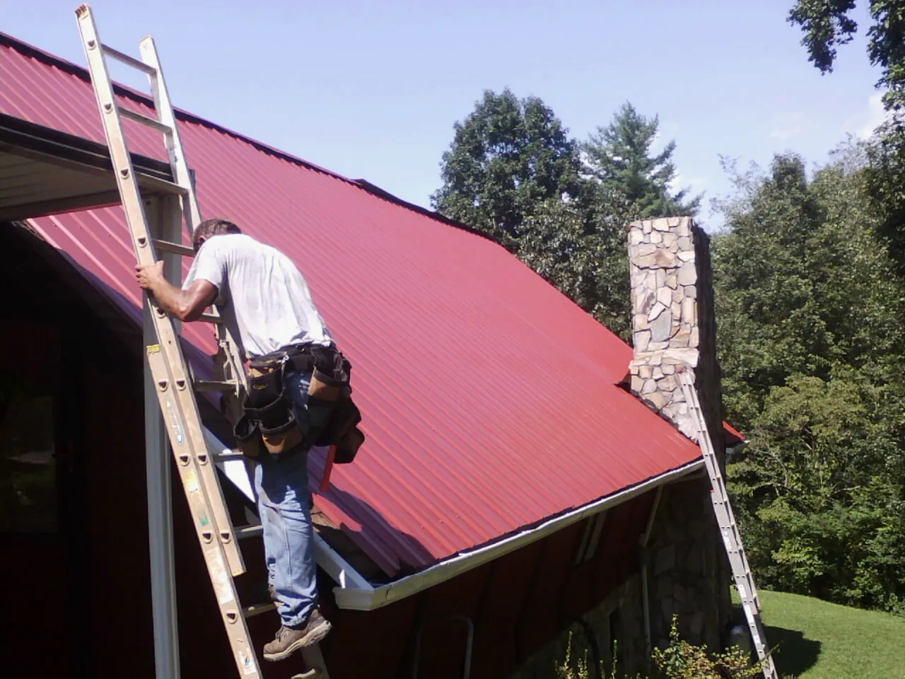 Roofer on a ladder beside a red metal roof and stone chimney — Soffit & Fascia Repair by Roofing Brothers in Malinta